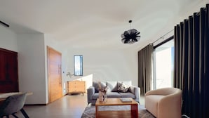 Uma sala de estar moderna com sofá, mesa de centro e poltrona. Há um armário de madeira, uma escrivaninha e uma janela com cortinas.