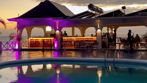 Ein Poolbereich mit Bar und Sitzgelegenheiten, umgeben von einer überdachten Struktur mit Bögen, mit Blick auf das Meer bei Sonnenuntergang.