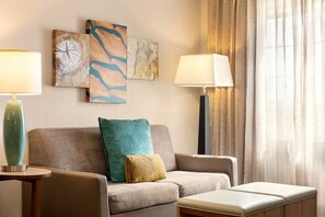 Un salon moderne avec un canapé, deux tables d’appoint et une lampe. Les murs sont ornés de tableaux abstraits et il y a une fenêtre avec des rideaux.