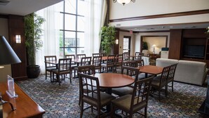 A hotel lobby with round tables and chairs, a sofa, potted plants, and a television.