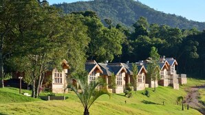 Uma fileira de casas de pedra com telhados inclinados, rodeada por vegetação exuberante e colinas ao fundo.