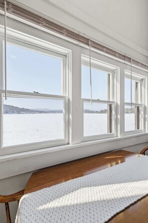 Una habitación con una mesa de madera y un tapete blanco de punto. Hay tres ventanas con marcos blancos, y se ve un paisaje nevado desde el exterior.