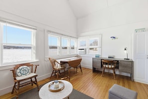 Habitación con piso de madera, paredes blancas y ventanas amplias con vista a la playa. Hay dos sillas de madera, una mesita redonda y un escritorio con silla.