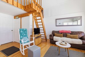 A cozy living room with a wooden staircase, a television on a wooden stand, a brown leather sofa, and a white door.