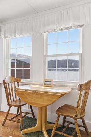 Un coin repas avec une table en bois et deux chaises, un tableau encadré posé sur la table, et deux fenêtres donnant sur un paysage enneigé.