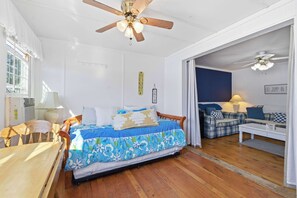 Un dormitorio con cama, suelo de madera, ventilador de techo y vistas a otra habitación con un sofá y una lámpara.