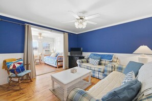 Una sala de estar con pared azul, piso de madera, ventilador de techo blanco, televisor, sofá, sillón mecedora y una mesa de centro.