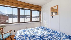 A bedroom with a large window, a bed with a blue and white patterned bedspread, a wooden rocking chair, and a ceiling fan.