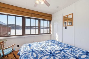 Une chambre avec une grande fenêtre, un lit recouvert d’une courtepointe à motifs bleus et blancs, un fauteuil à bascule en bois et un ventilateur de plafond.