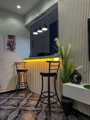 Eine moderne Bar mit zwei schwarzen Barhocker, einer Theke mit gelber Beleuchtung und einer dekorativen Vase auf einer weißen Konsole.