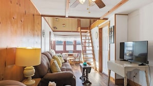 A cozy living room with a brown leather sofa, a white desk with a television, and a wooden ceiling fan.