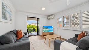 Uma sala de estar moderna com sofás cinzas, uma mesa de centro de madeira e uma TV em um móvel. Há uma varanda com mobiliário externo e vista para edifícios.