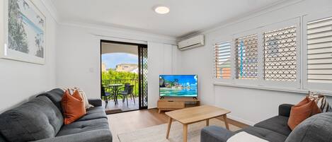A modern living room with grey sofas, a wooden coffee table, and a TV on a stand. There is a balcony with outdoor furniture and a view of buildings.
