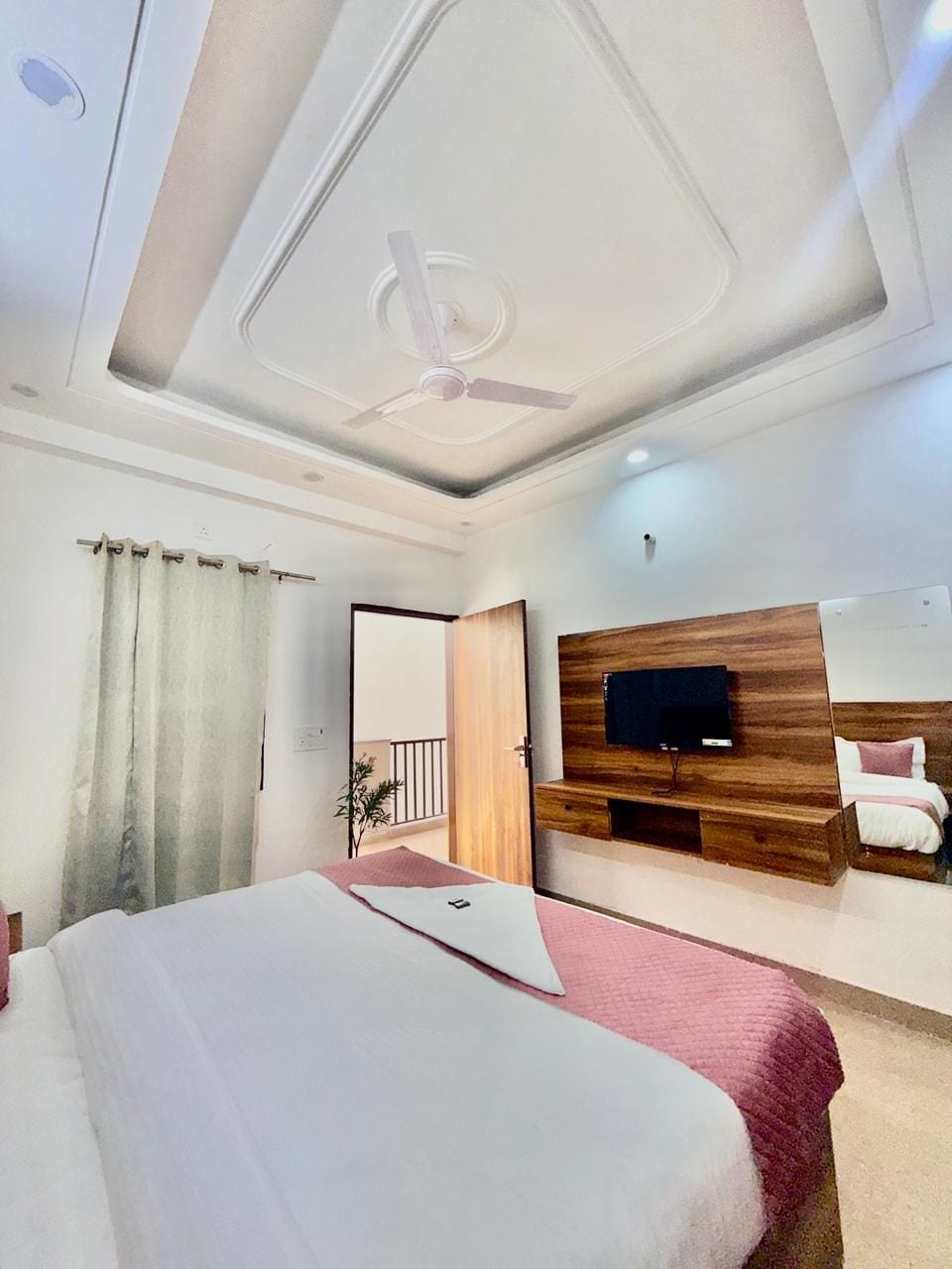 A modern hotel room with a bed, a TV mounted on a wooden panel, a ceiling fan, and a view of the outside balcony.