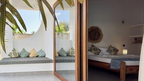 Una habitación con cama, un espejo, un estante en la pared y una ventana con vista a follaje.