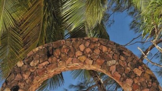 Un arco de piedra con una hamaca colgando en su interior, con vistas a una playa de aguas turquesas cristalinas y palmeras.