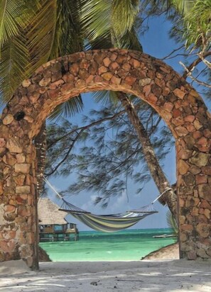 Un arco de piedra con una hamaca colgando en su interior, con vistas a una playa de aguas turquesas cristalinas y palmeras.