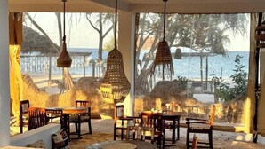 Un área de comedor frente a la playa con luces colgantes de mimbre, mobiliario de madera y vistas al mar y palmeras.