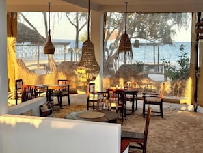 Un área de comedor frente a la playa con luces colgantes de mimbre, mobiliario de madera y vistas al mar y palmeras.