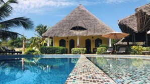 Una piscina con borde de azulejos, un edificio con techo de paja y palmeras.