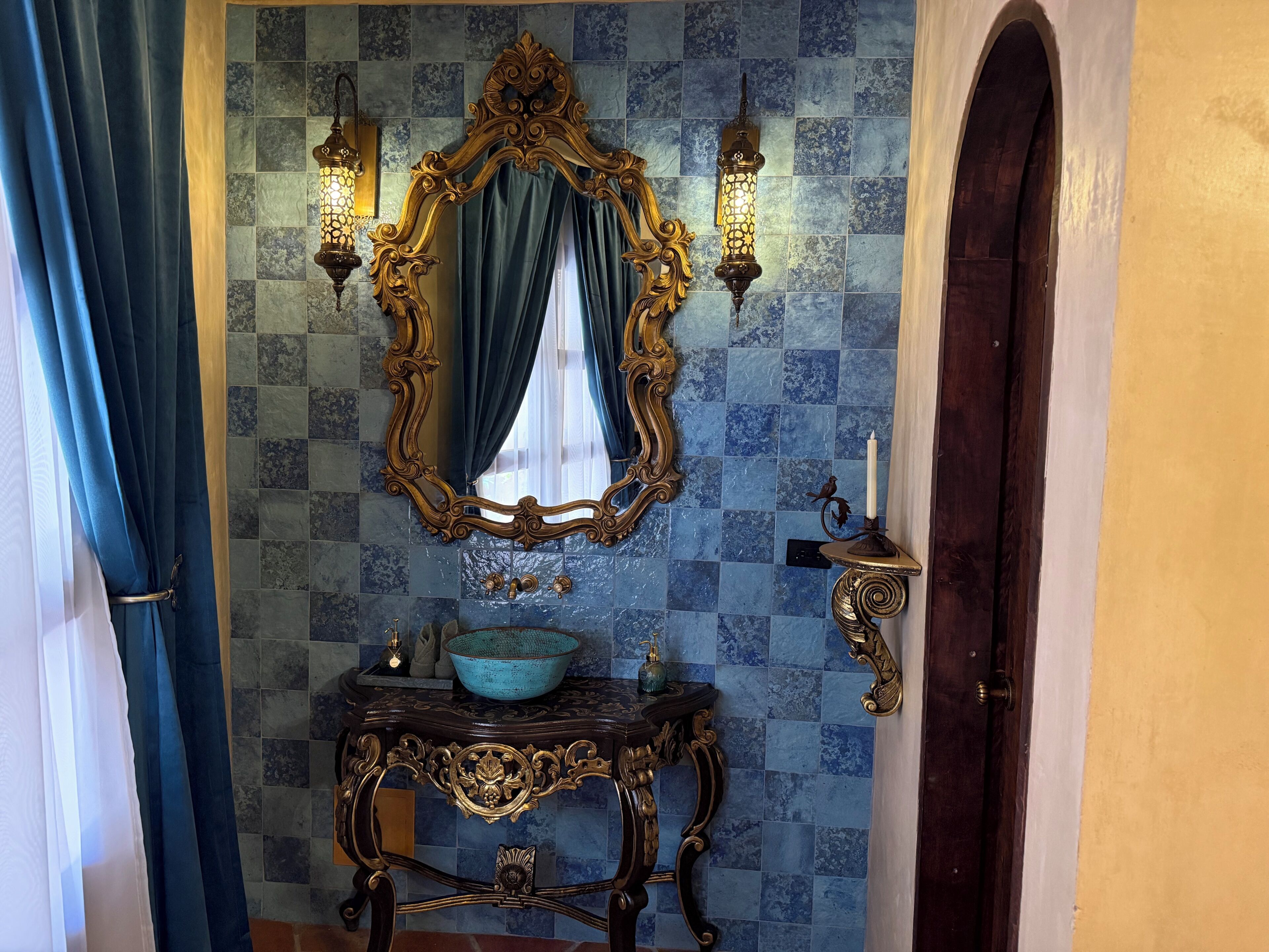 Um banheiro com um espelho decorativo, luminárias embutidas na parede, uma pia com cuba azul e uma porta.