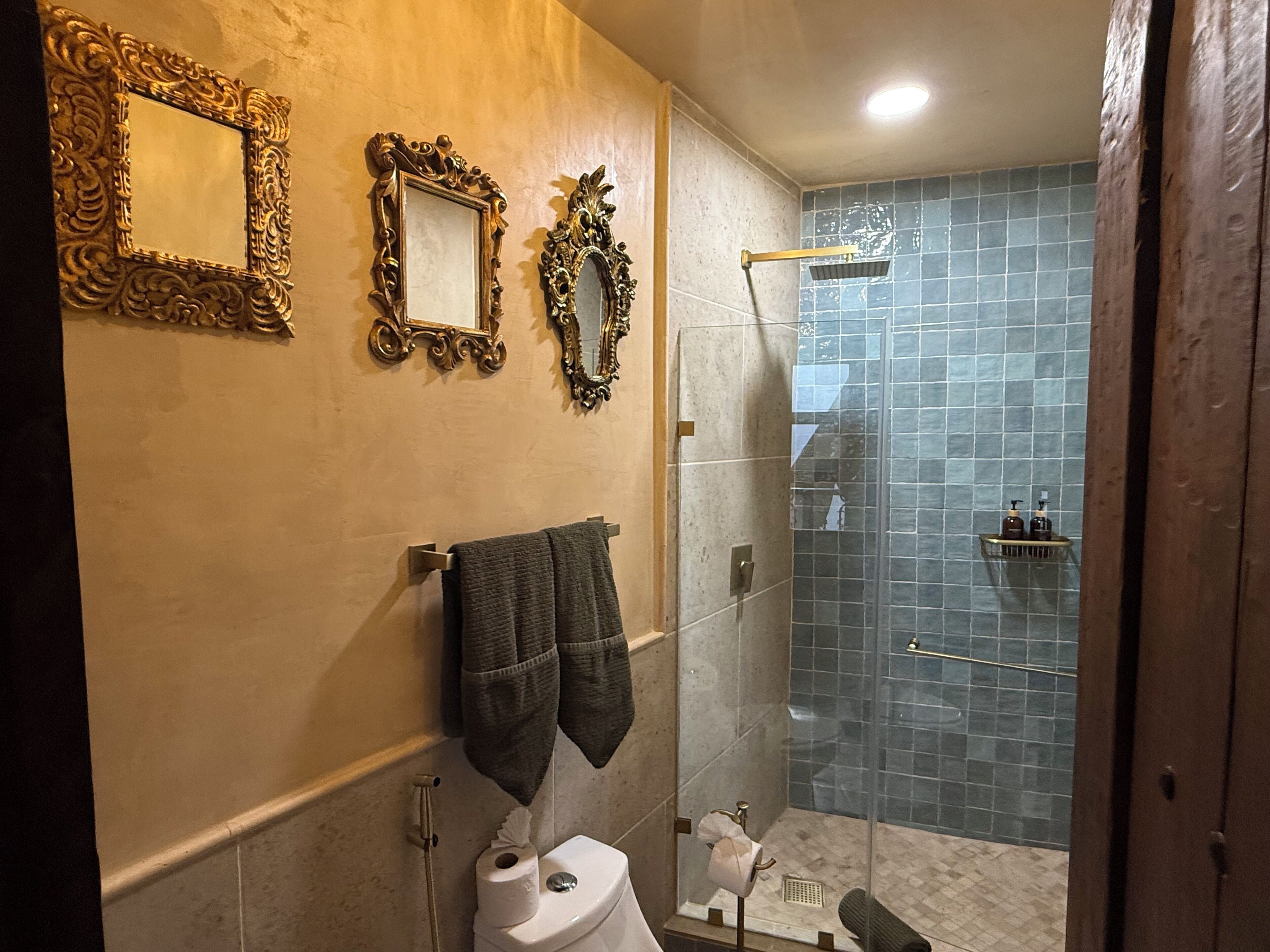 A bathroom with a toilet, a towel rack with a towel, a shower with a glass door, and three ornate mirrors on the wall.