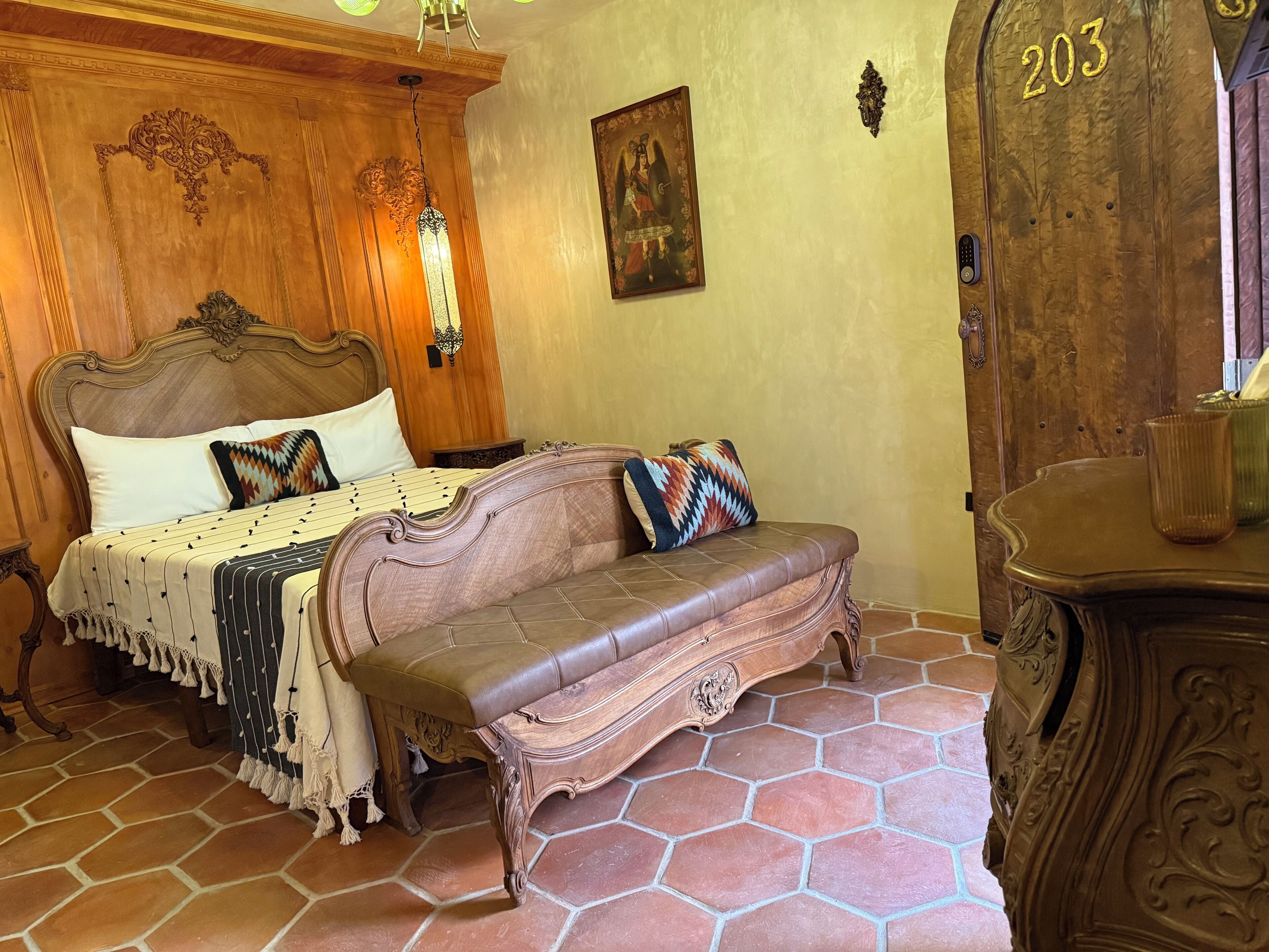 Un dormitorio con cabecera de madera, cama con almohadas estampadas, una banca y una puerta con un número.