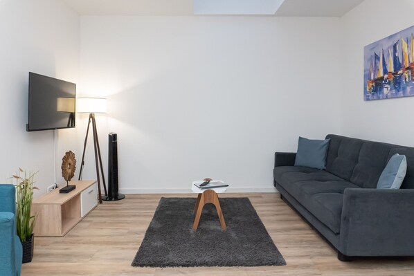 A modern living room with a grey sofa, a small wooden table, a floor lamp, a TV mounted on the wall, and a painting. - Apartmenthaus am Park Wohnung 1 (Wilhelmshaven)