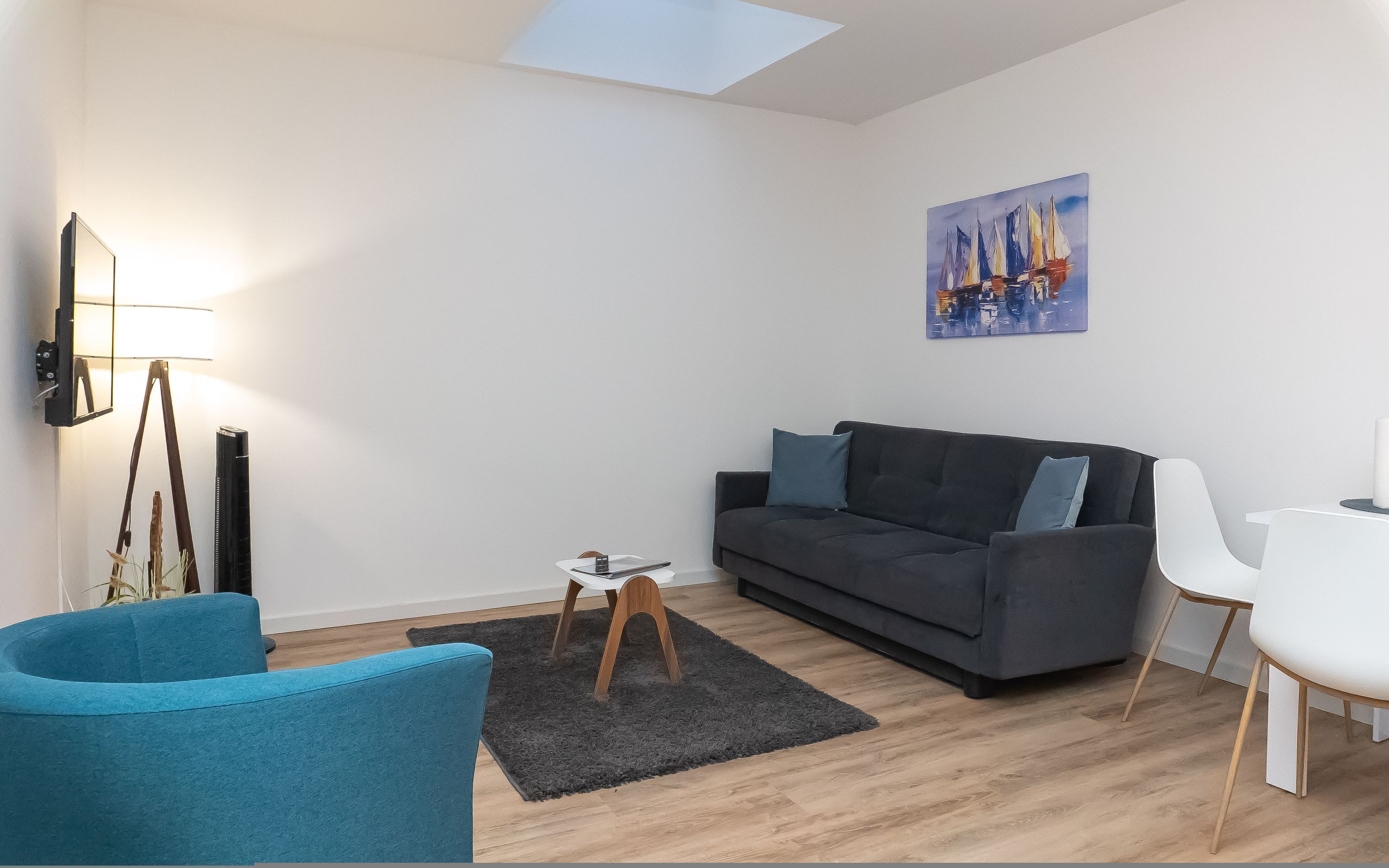 Una sala de estar moderna con un sofá gris, un sillón azul, una mesita de centro y un cuadro de veleros en la pared.