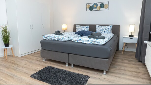 A modern bedroom with a grey sofa bed, a small bedside table with a lamp, a potted plant, and a wall art piece. - Apartmenthaus am Park Wohnung 1 (Wilhelmshaven)