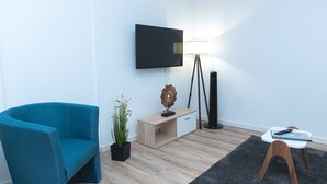 A modern living room with a teal armchair, a wooden side table, a floor lamp, a TV mounted on the wall, and a small table with a remote control. - Apartmenthaus am Park Wohnung 1 (Wilhelmshaven)