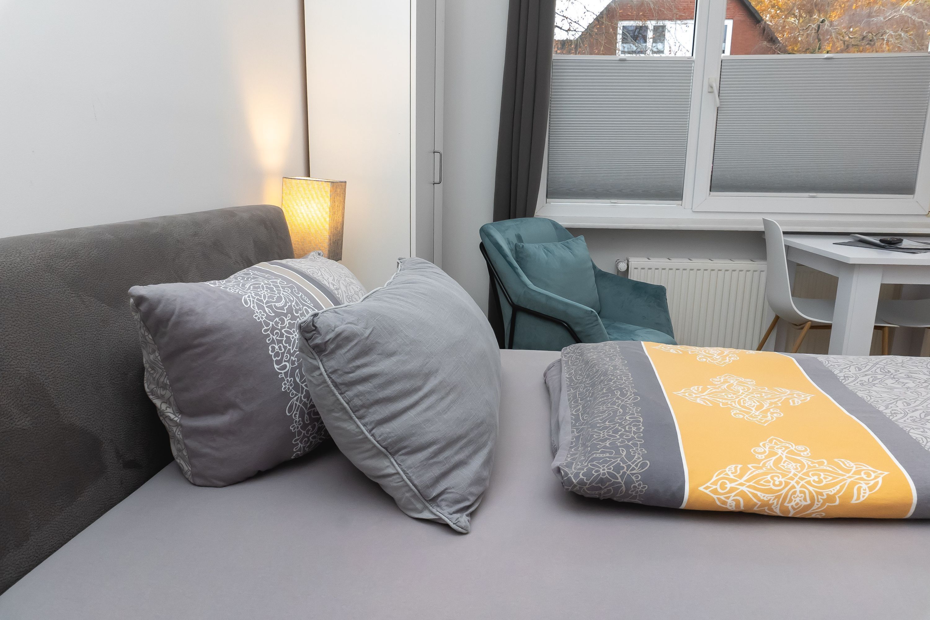 A bedroom with a bed, two pillows, a lamp, a chair, and a window with blinds.