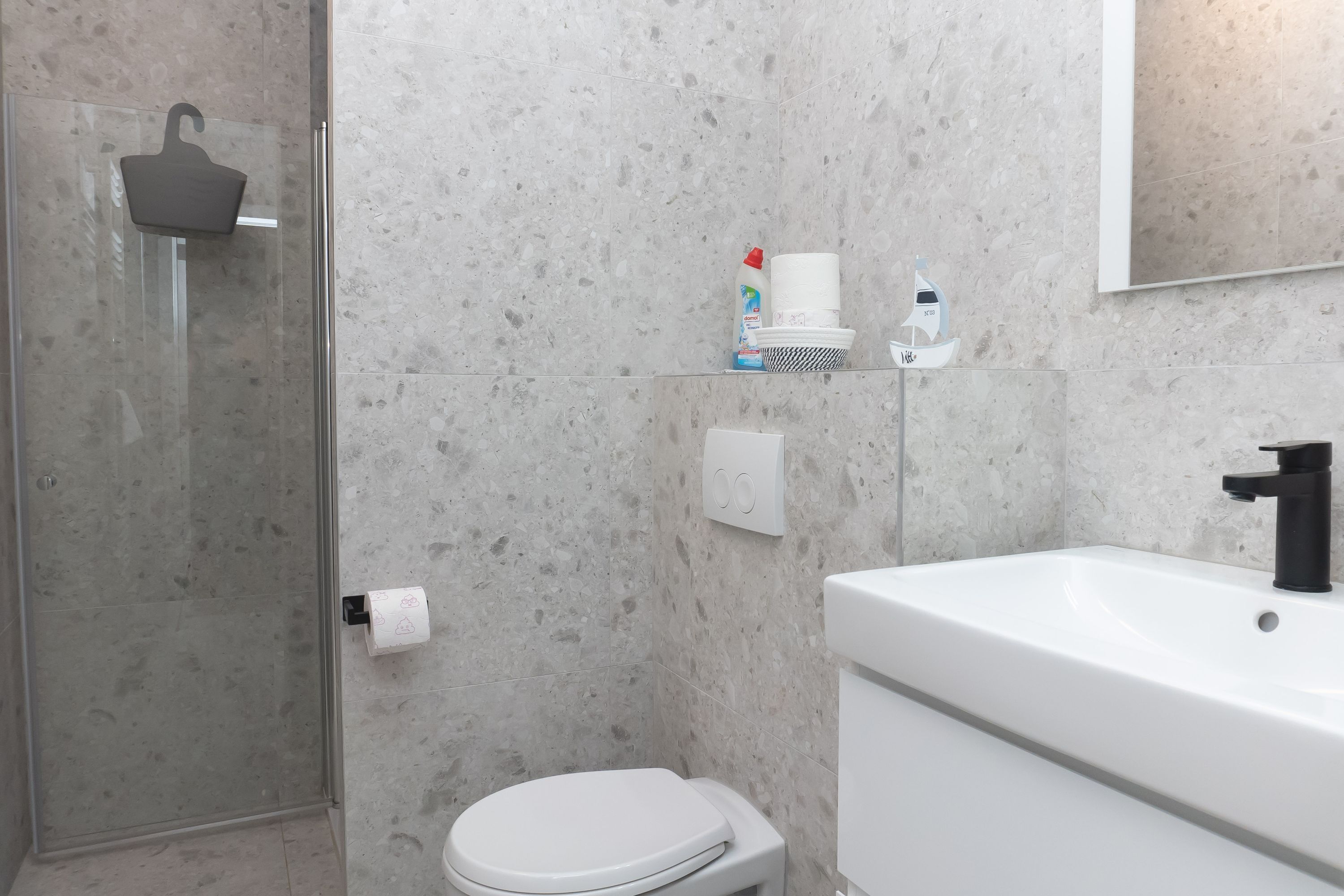 A modern bathroom with a glass shower enclosure, a toilet, a sink with a black faucet, and toiletries on a shelf.