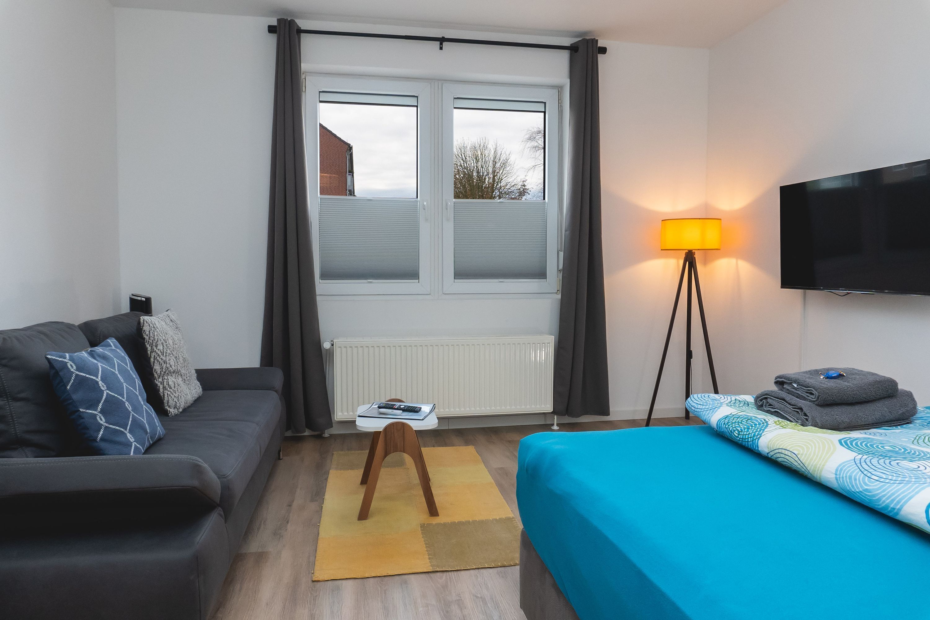 A modern hotel room with a grey sofa, a blue bed, a standing lamp, and a TV on the wall.