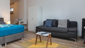 Una stanza moderna e luminosa con un divano grigio, un piccolo tavolo bianco e un letto con copriletto blu.