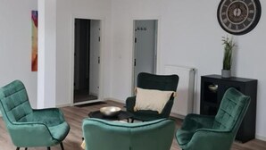 A modern living room with green upholstered chairs, a black sofa, and a round coffee table. There are two pendant lights with a copper finish. A decorative clock is on the wall, and a plant is on a black side table. - Apartmenthaus am Park Wohnung 7 (Wilhelmshaven)