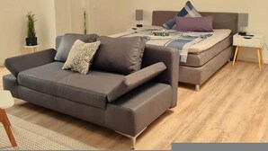 A modern bedroom with a sofa, a bed with pillows, a nightstand, a lamp, and a painting on the wall. - Apartmenthaus am Park Wohnung 7 (Wilhelmshaven)
