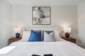 Una cama bien hecha con ropa blanca y almohadas azules, un cuadro abstracto grande encima y dos lámparas de noche sobre mesitas de madera.