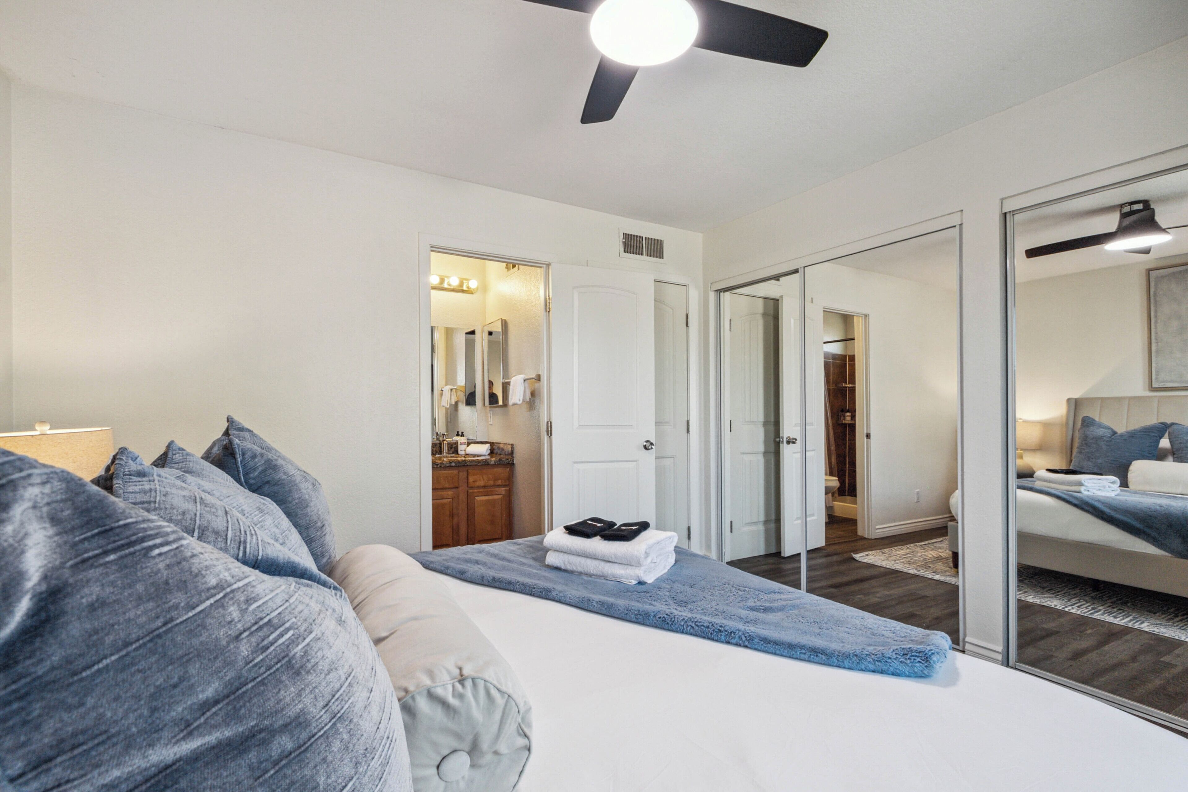 Un dormitorio con cama, ventilador de techo, baño con espejo y armario.