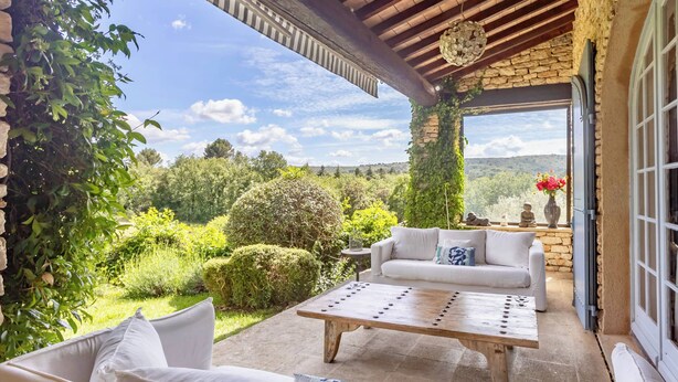 A patio with a stone table, white sofas, and a view of a lush garden and hills.