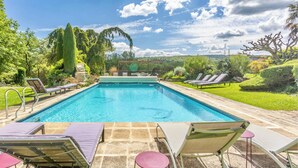 A rectangular swimming pool with clear blue water, surrounded by a stone deck. There are lounge chairs and a covered seating area with a pergola.