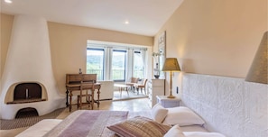 Une chambre avec une cheminée, un lit avec des oreillers, une table de chevet avec une lampe, une commode et une vue sur l’extérieur par une fenêtre.