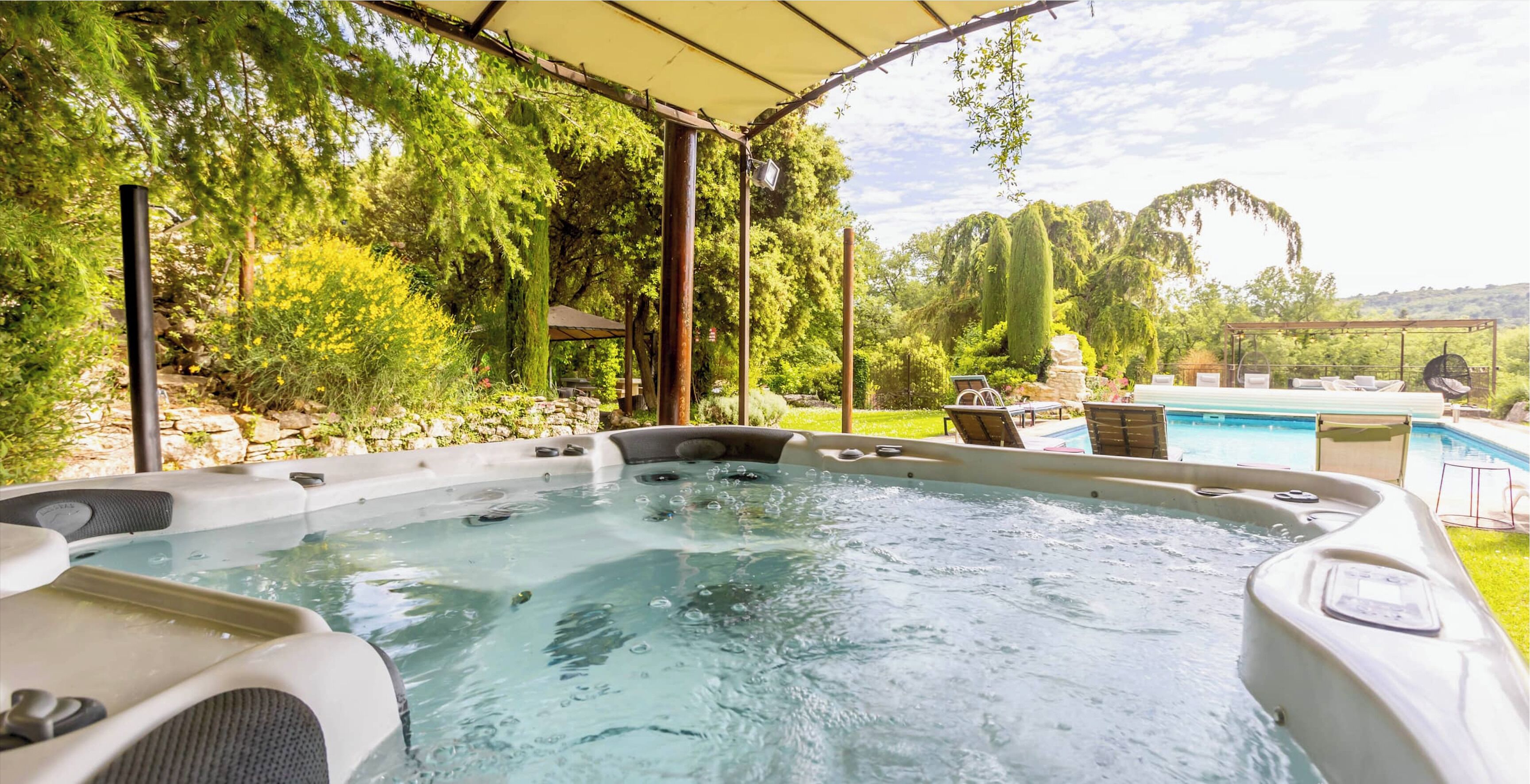 Un espace avec un jacuzzi, agrémenté d’une pergola couverte, entouré d’une végétation luxuriante et d’une piscine.