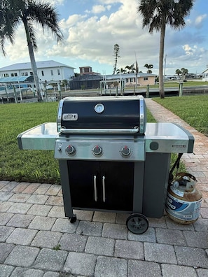 A portable Weber Genesis II grill with a gas tank, located on a paved area with grass and palm trees in the background. - NEW POOL+ 3 Bedroom + No Bridge Gulf Access + Dock (Englewood)