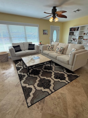 A living room with a sectional sofa, a coffee table, a patterned rug, a ceiling fan, and a bookshelf. - NEW POOL+ 3 Bedroom + No Bridge Gulf Access + Dock (Englewood)