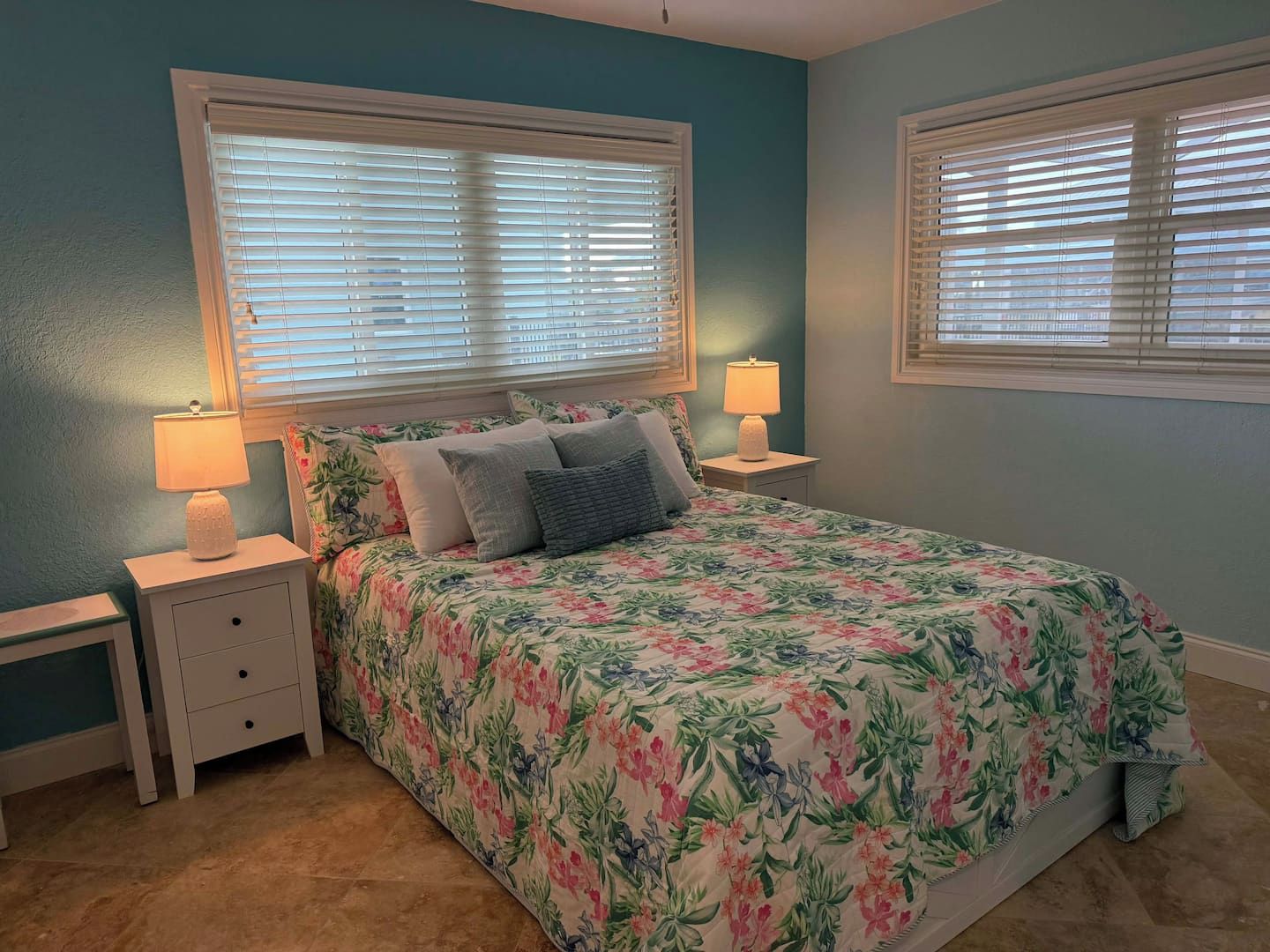 Un dormitorio con edredón estampado de flores, dos mesitas de noche con lámparas y una ventana con persianas.