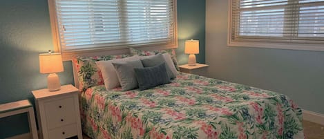 A bedroom with a floral bedspread, two bedside tables with lamps, and a window with blinds.