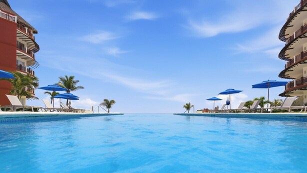 Une piscine d’un bleu éclatant, bordée de chaises longues et d’ombrelles, et entourée de palmiers et de bâtiments d’hôtel.