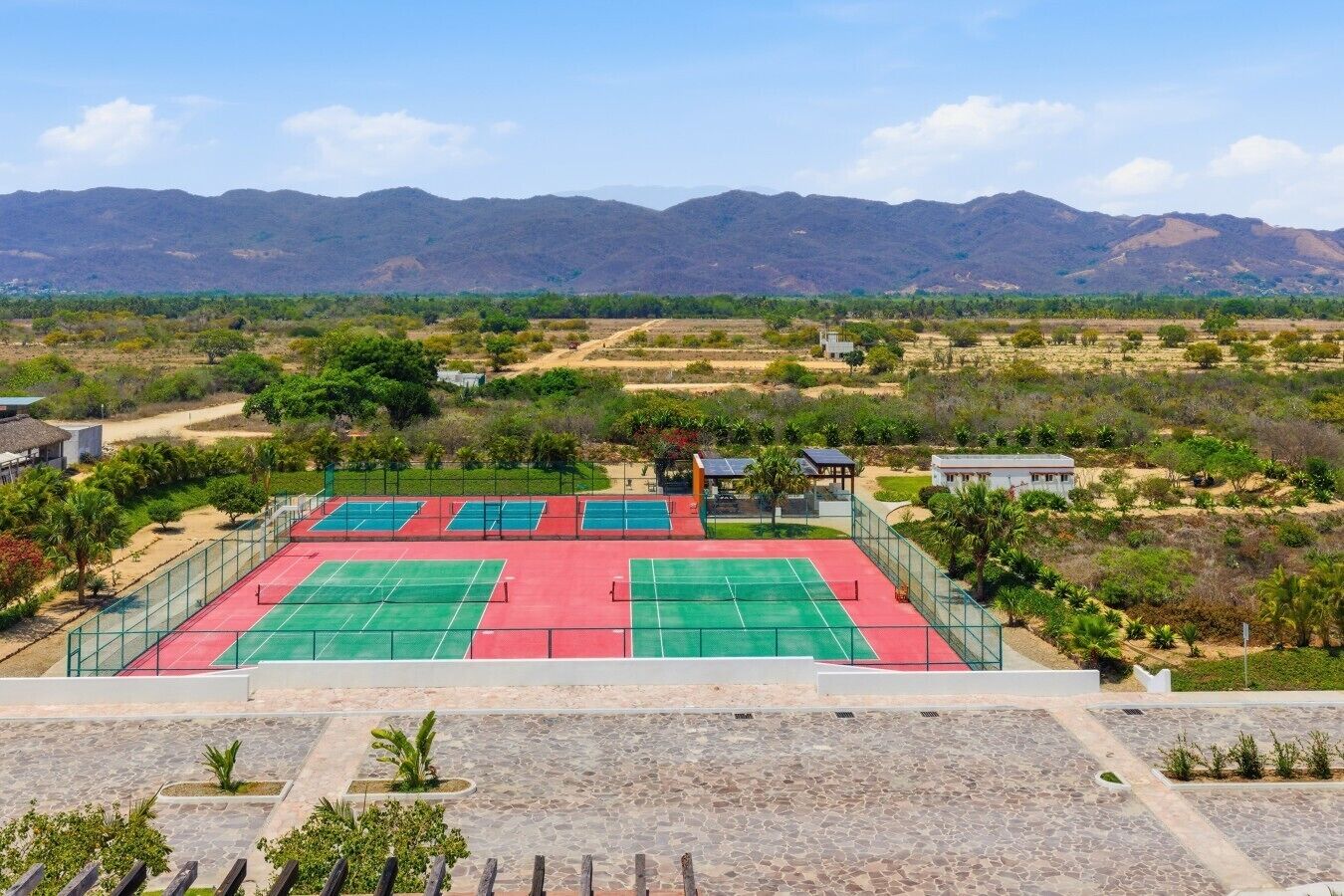 A tennis court complex with multiple courts, surrounded by greenery and mountains in the background.