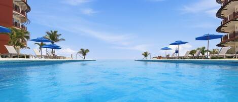 A clear blue pool with lounge chairs and umbrellas, surrounded by palm trees and hotel buildings.